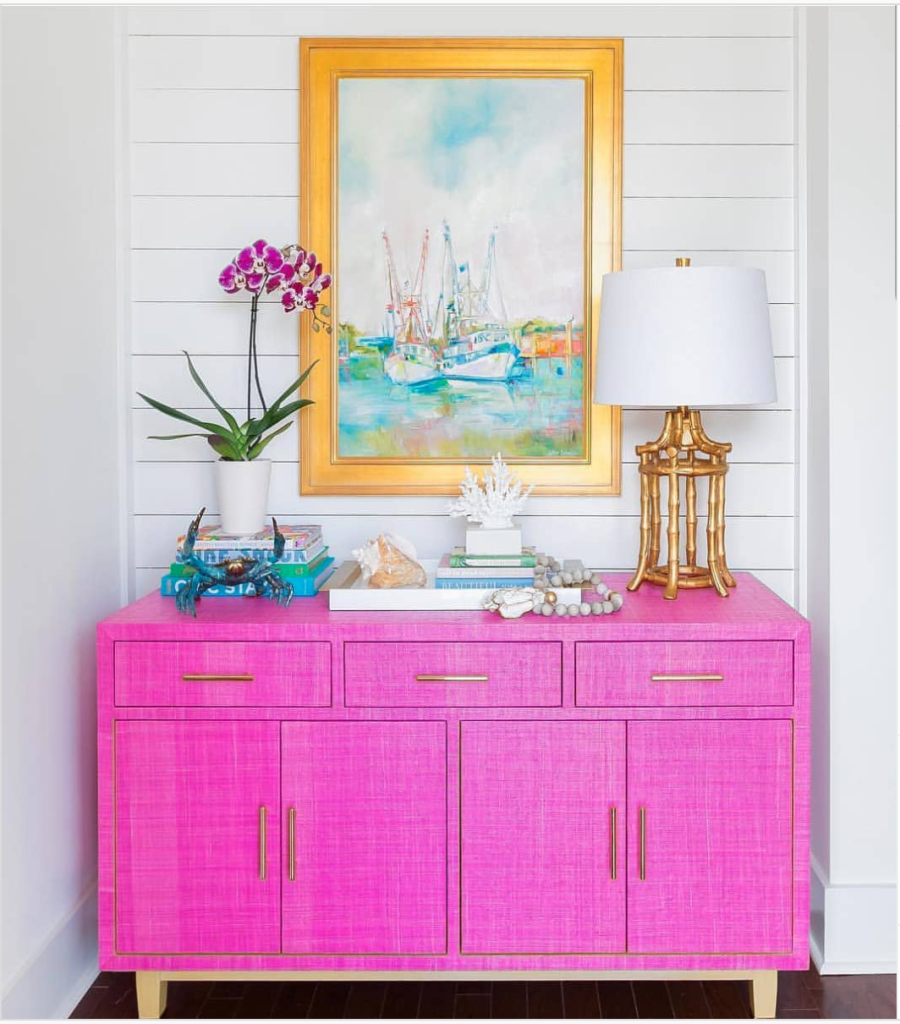 A pink credenza in an entryway with a painting of fishing boats above it with extra accessories